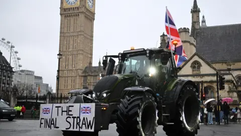 Getty Images A farmer waves a Union flag from their cab as they drive a tractor past the Houses of Parliament, during a protest against inheritance tax rules for land ownership, in London. Big Ben is in the background.